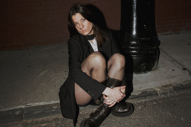 Promotional portrait of Kate "Babyshakes" Fuller of Full Flower Moon Band sitting on a sidewalk at night.
